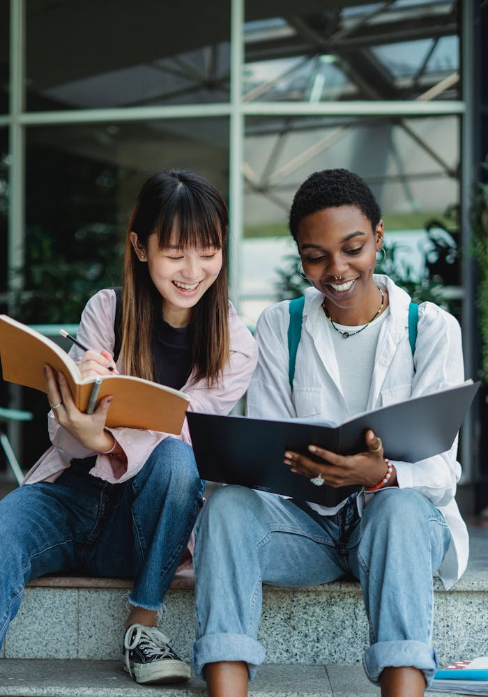 Smiling young multiracial ladies sitting outside in casual outfit with notebooks and talking with each other in daytime