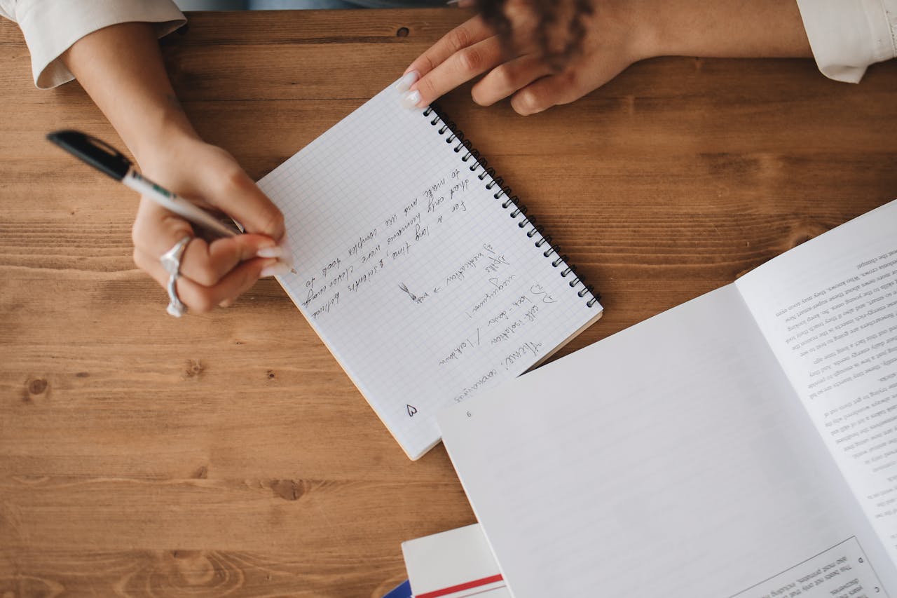Close-up view of a person taking notes in a notebook with an open book on a wooden desk.