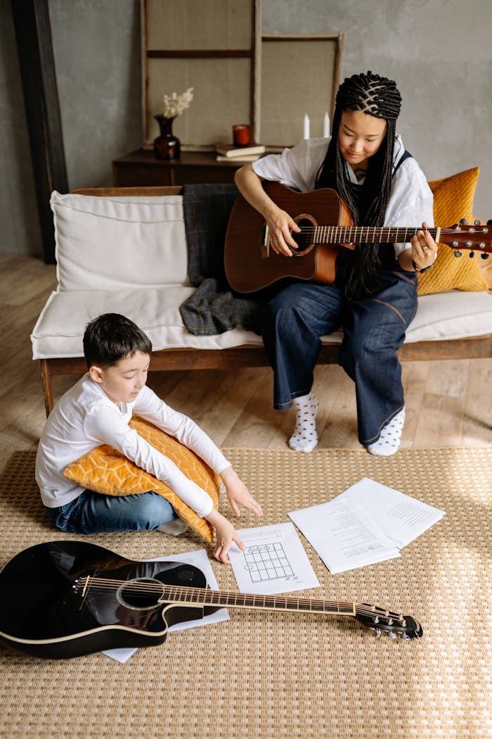 Child learning guitar at home with a female teacher, sitting on the floor with music sheets.