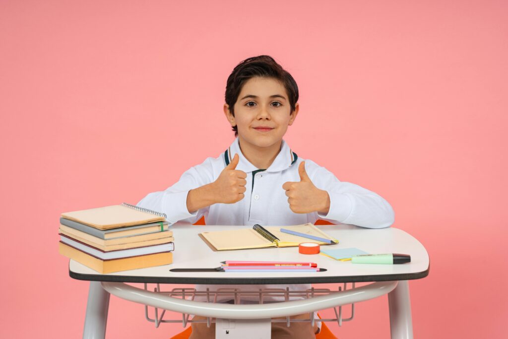 A happy schoolboy giving thumbs up while seated at a desk with school supplies, conveying positivity in education.