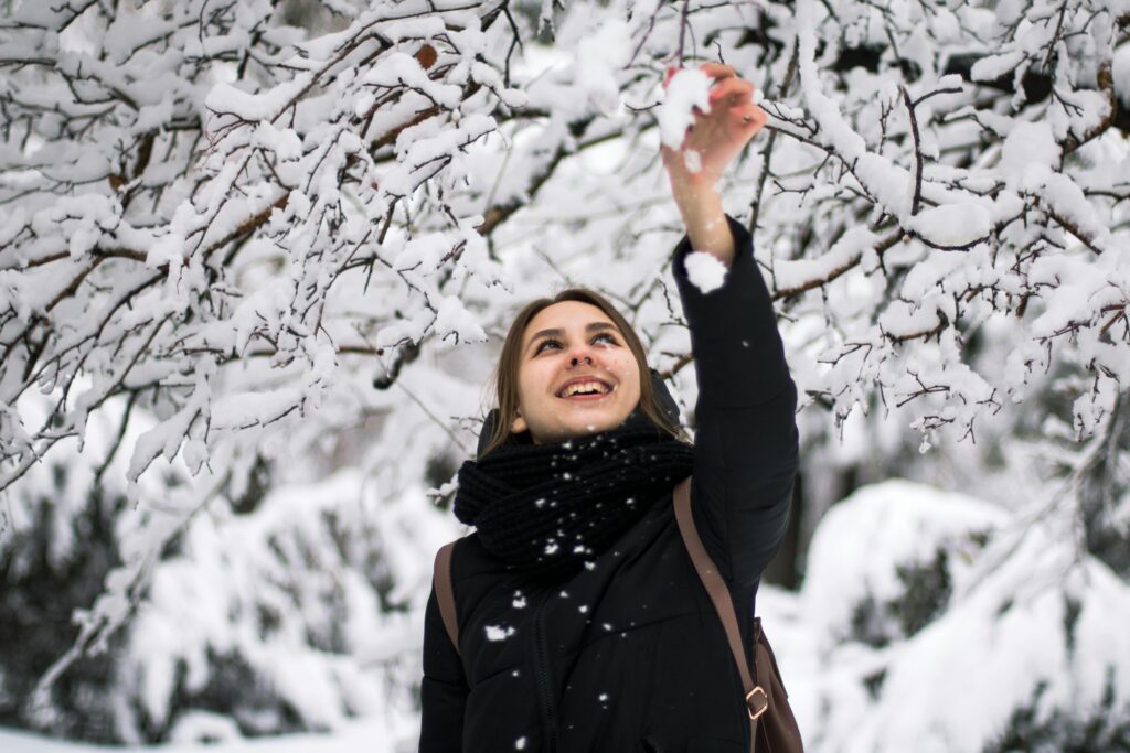 A joyful woman reaches for snow on tree branches in a winter wonderland, smiling warmly.