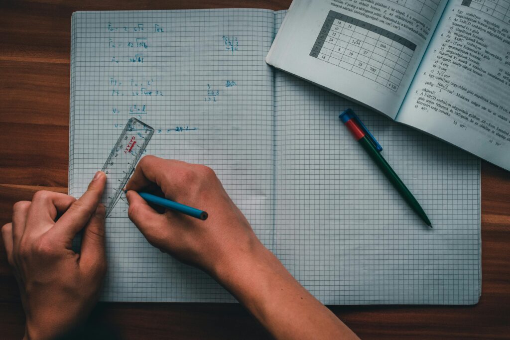 A person writes mathematical equations in a notebook using a ruler and pencil, focused on solving problems.