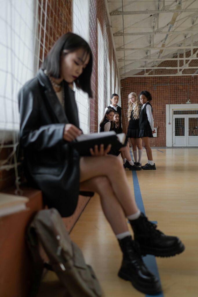 Teen girl reading alone in gymnasium while peers watch, highlighting themes of solitude and bullying.