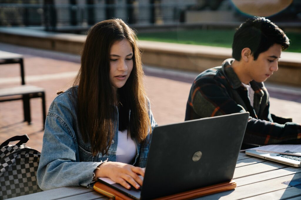 Two young adults working on laptops at an outdoor table.