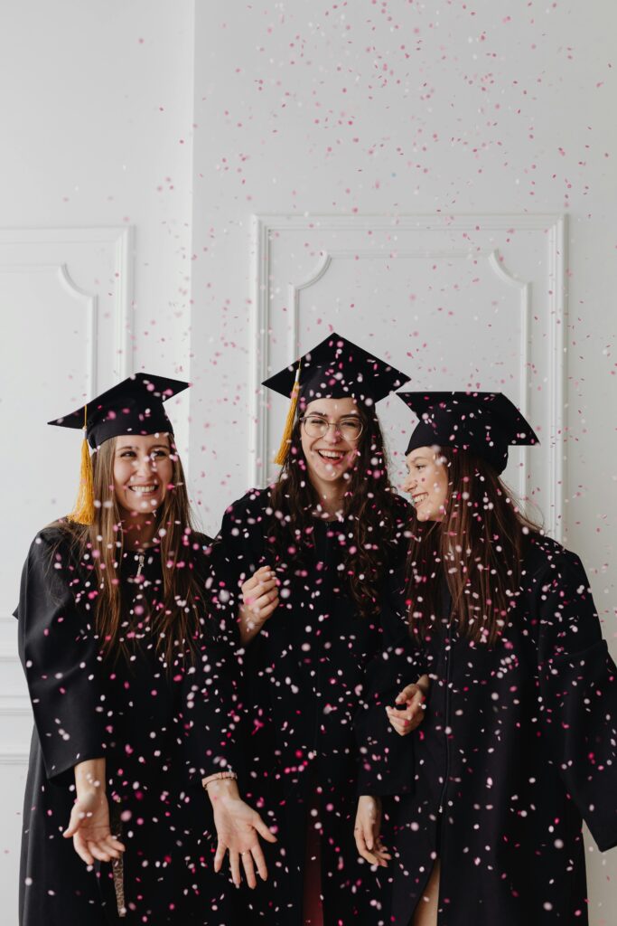 Three women graduates joyfully celebrate with confetti indoors, showcasing excitement and success.