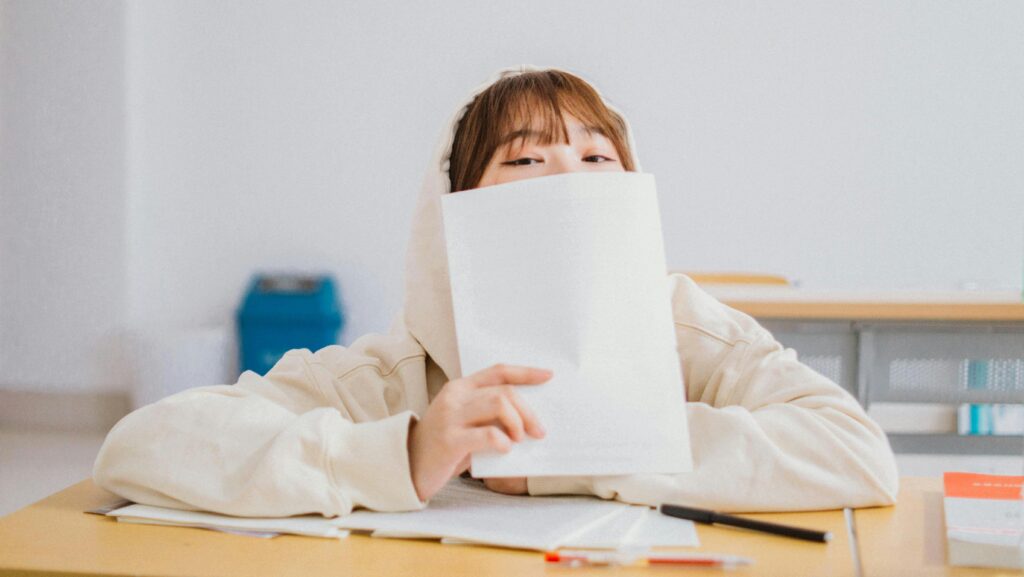 A young woman seated at a desk, partially covered by a sheet of paper in a classroom setting, conveying curiosity.