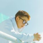 A young man in a white hoodie leaning on a railing under a clear blue sky.